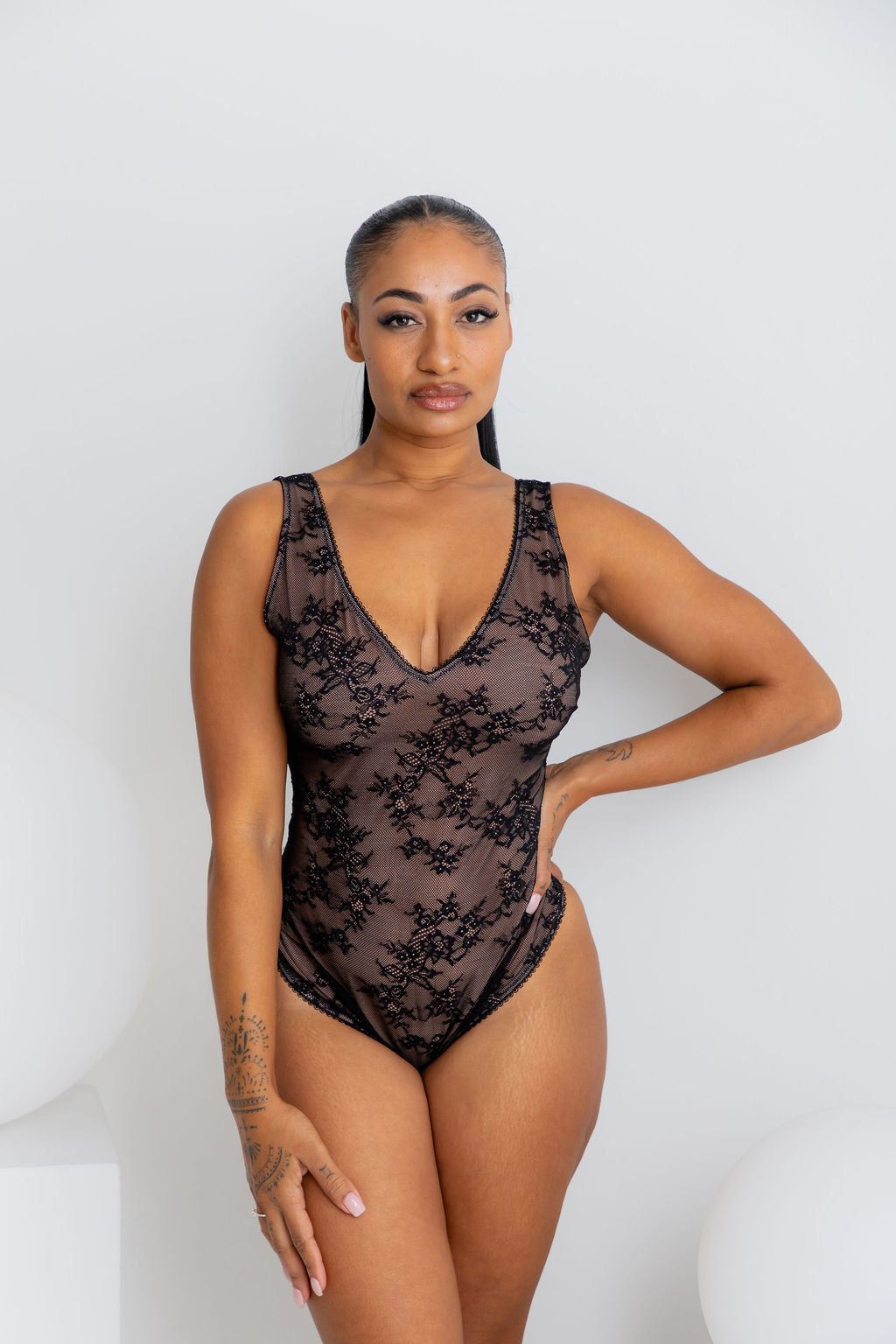 A woman modeling a black floral lace bodysuit with a deep V-neckline against a clean white studio background.