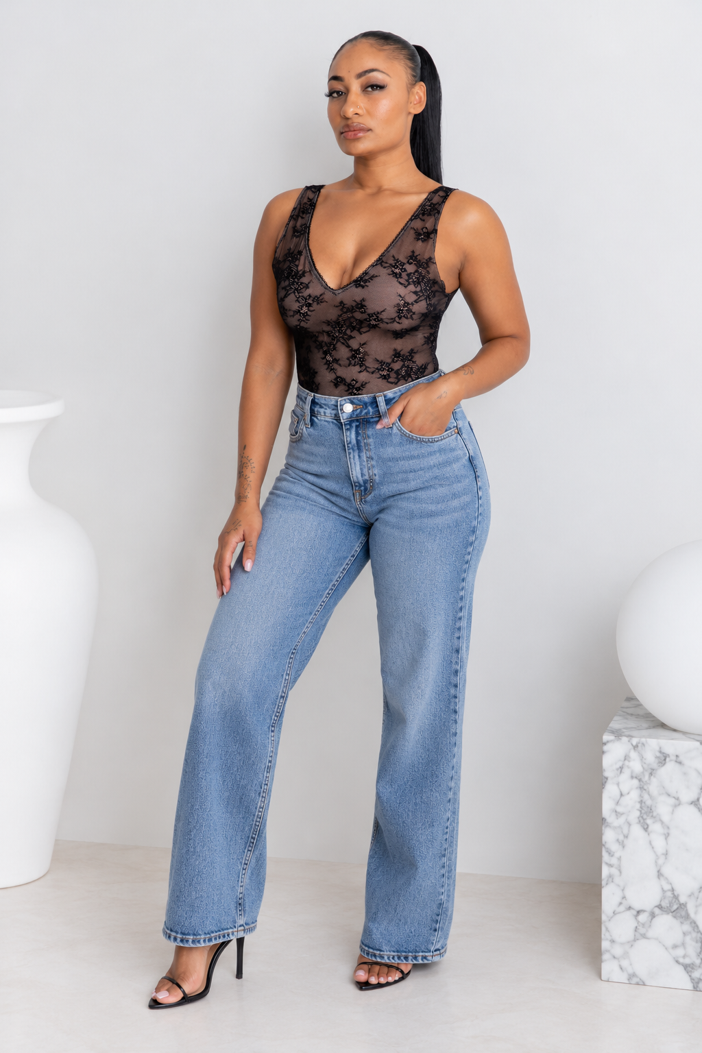 A woman modeling a black lace bodysuit with a deep V-neck and wide-leg blue jeans, standing in a minimalist studio setting with white decorative vases.