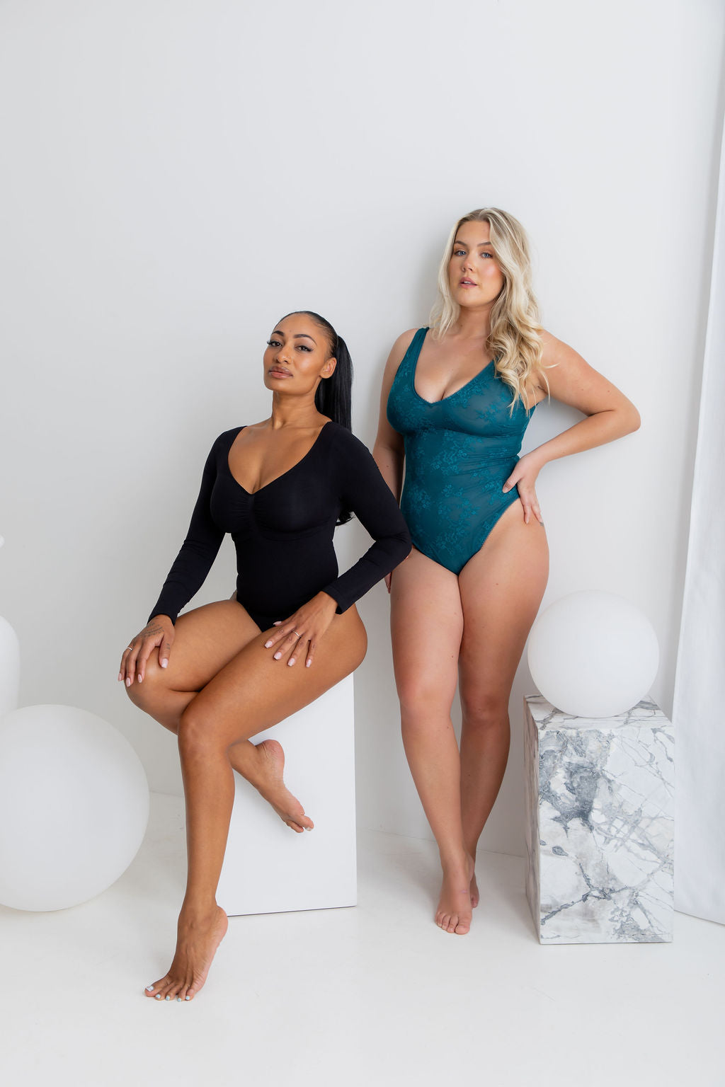 Two models posing in matching black and teal long-sleeve bodysuits against a white studio background with minimalist props.