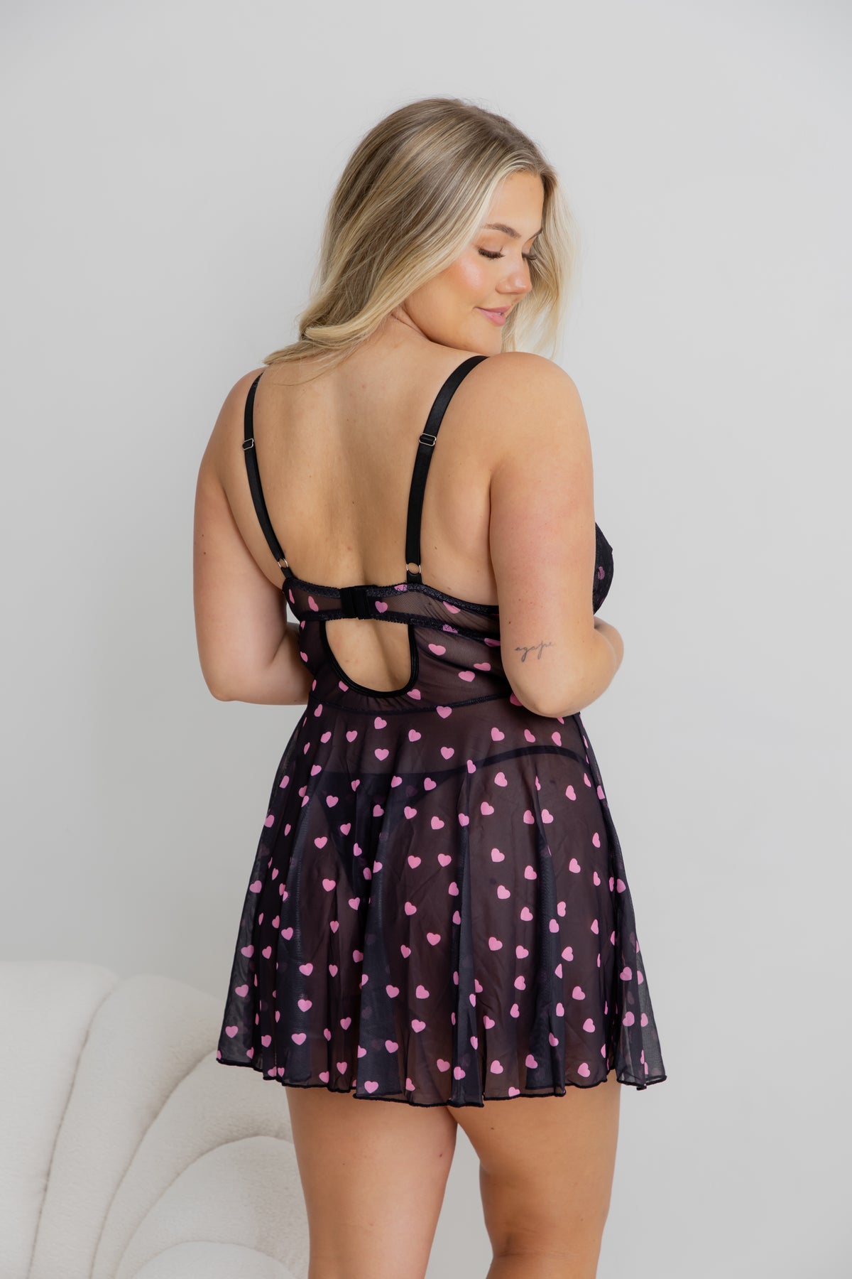 A woman wearing a sheer black babydoll lingerie dress with pink heart print, shown from the back against a light gray wall.