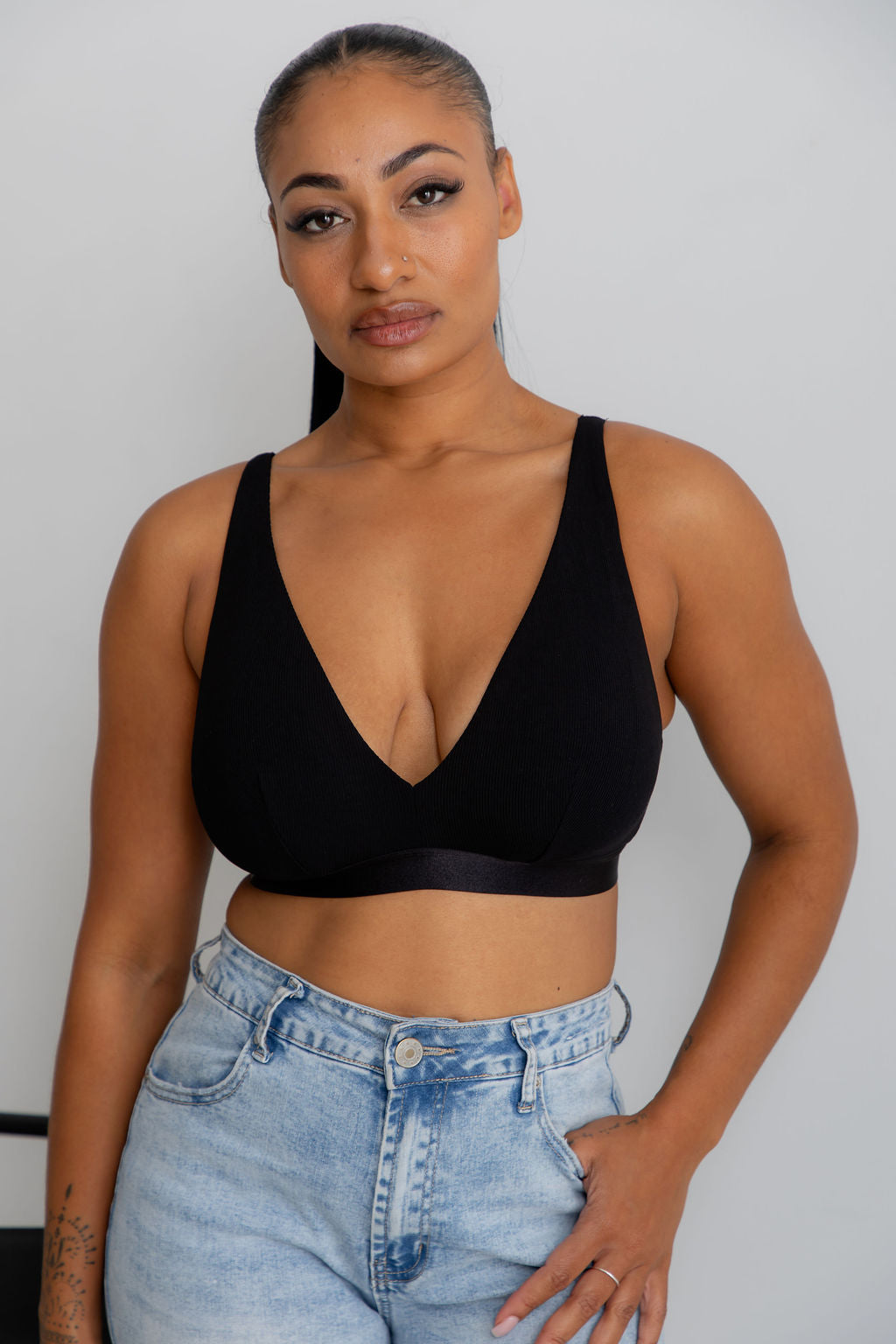 A woman modeling a black deep V-neck bralette with wide shoulder straps, paired with light-wash blue jeans, against a plain light gray wall.