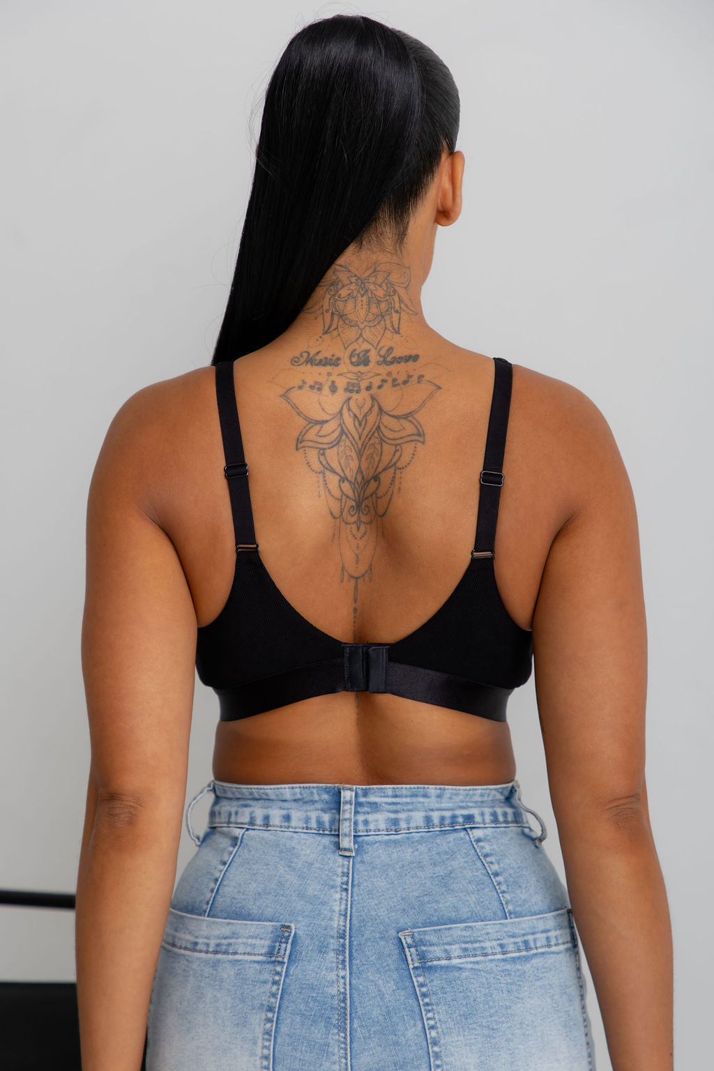 Rear view of a woman with long black hair and a large back tattoo wearing a black bralette and light blue denim shorts against a light gray background.