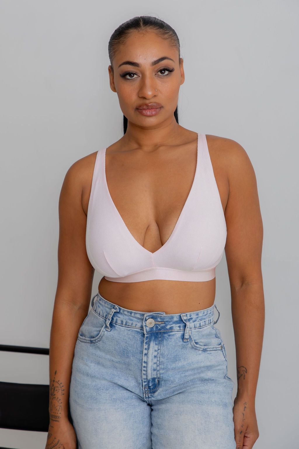 A woman wearing a light pink scoop-neck bralette and light-wash blue jeans, standing against a plain light gray wall.