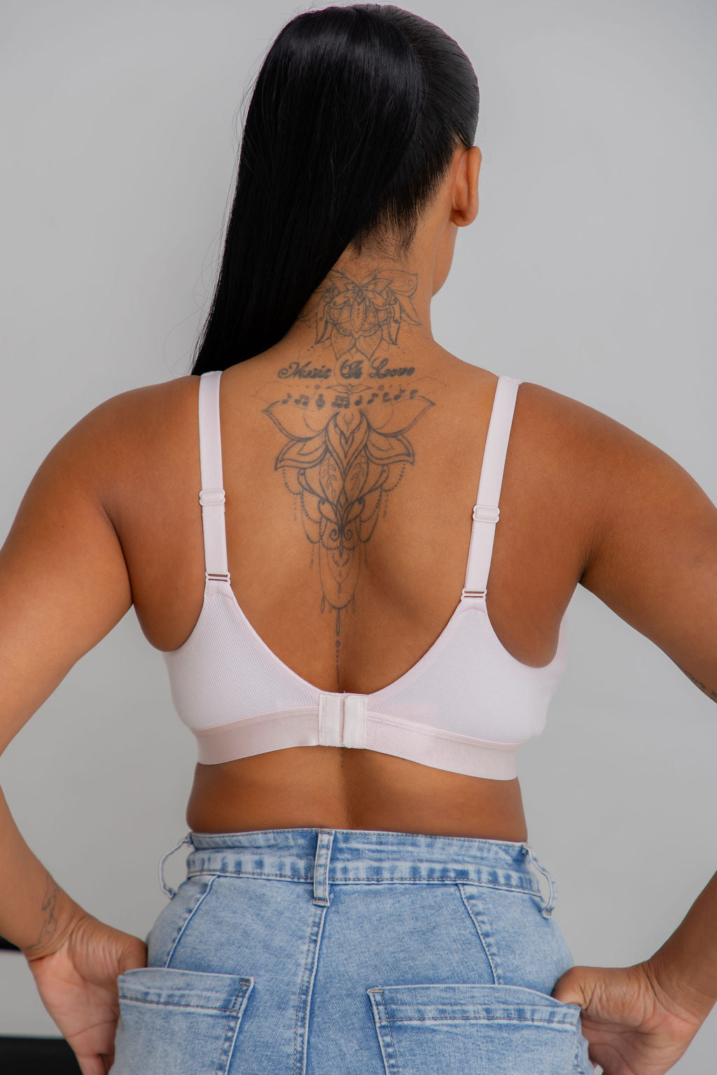 A woman viewed from the back wearing a pale pink underwire bra with thin adjustable straps and a center gore, paired with light blue denim jeans against a plain light gray background.