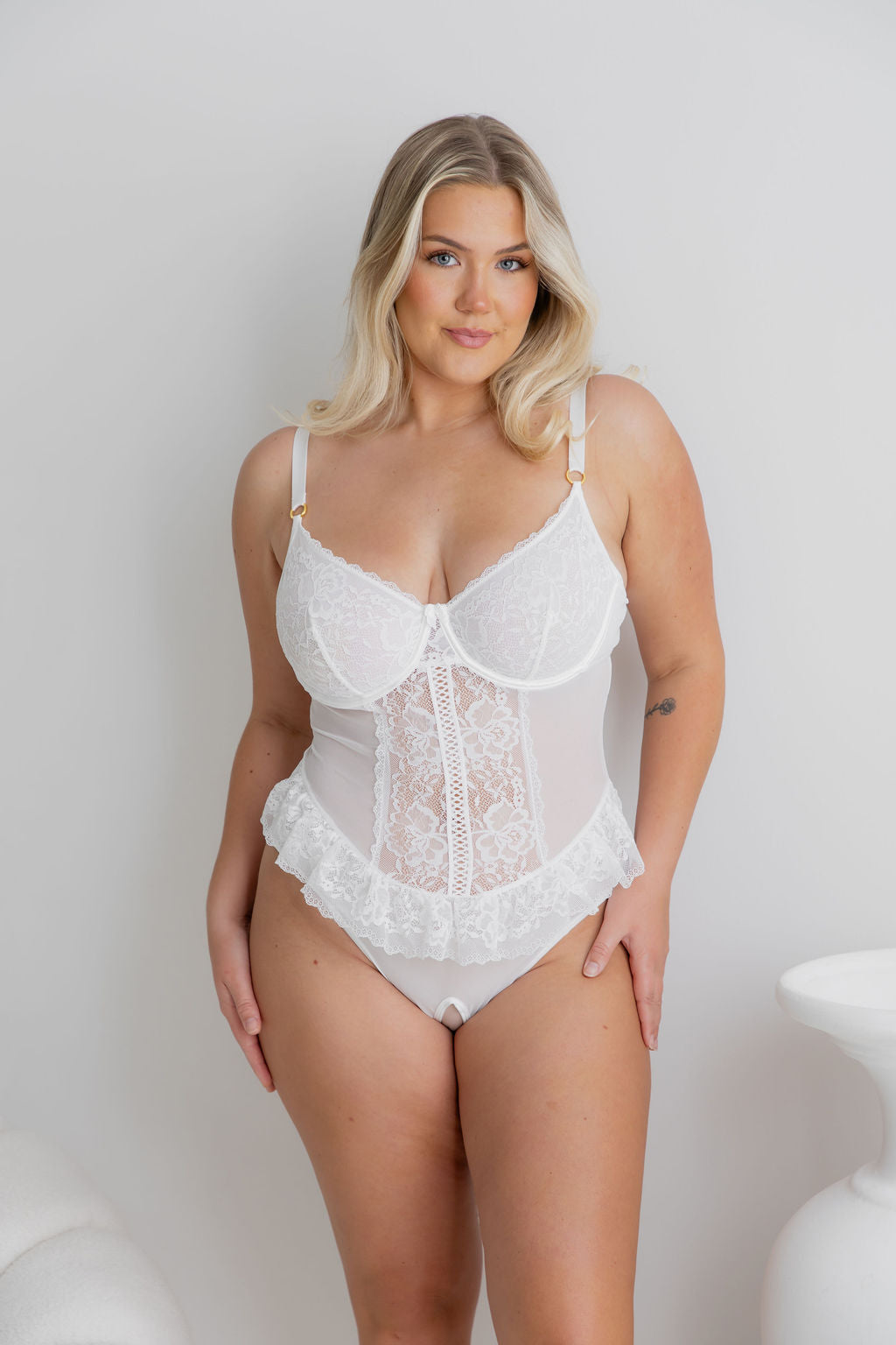 A blonde model wearing a white lace and mesh lingerie bodysuit with ruffled trim, standing against a light gray studio wall.