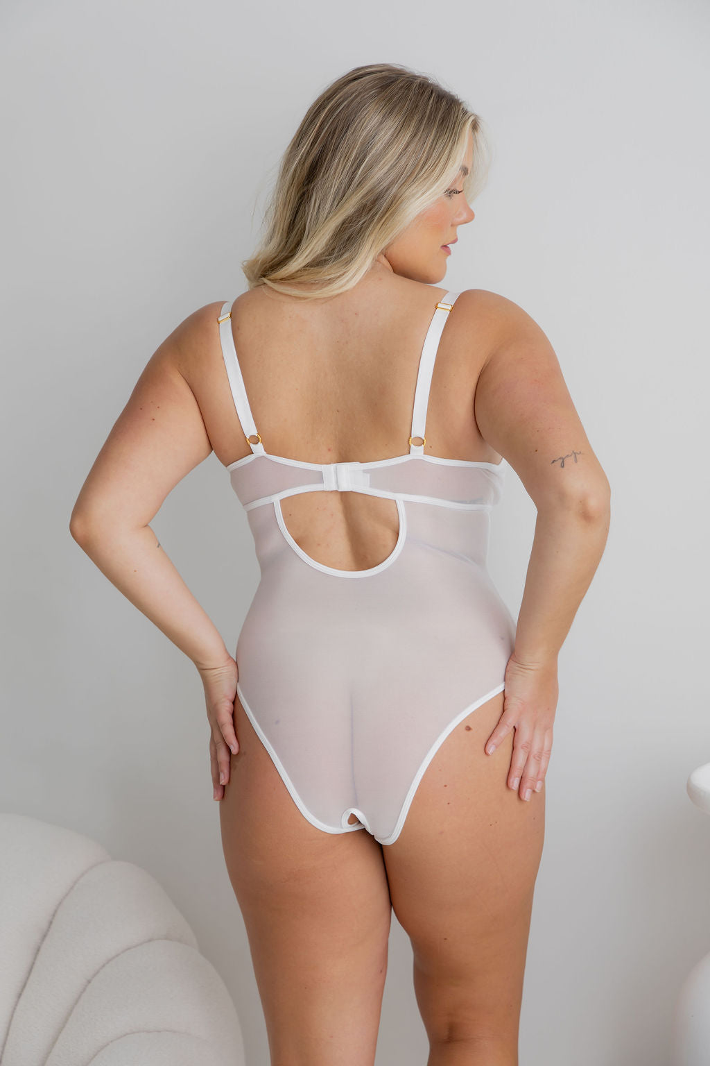 A woman wearing a light beige sheer bodysuit with white trim, shown from the back in a studio setting.
