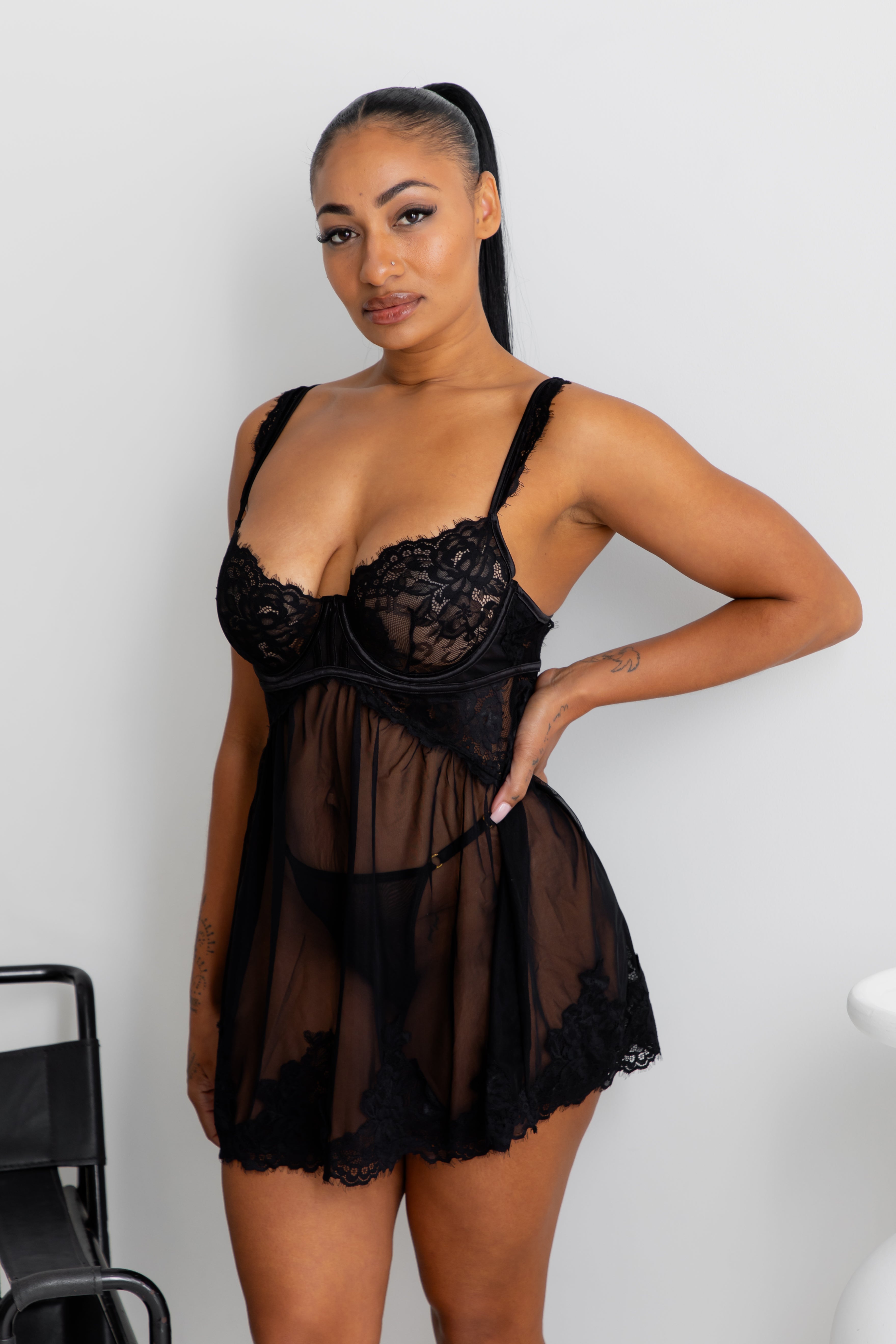 A woman modeling a black lace and sheer mesh babydoll lingerie chemise against a plain light gray wall.