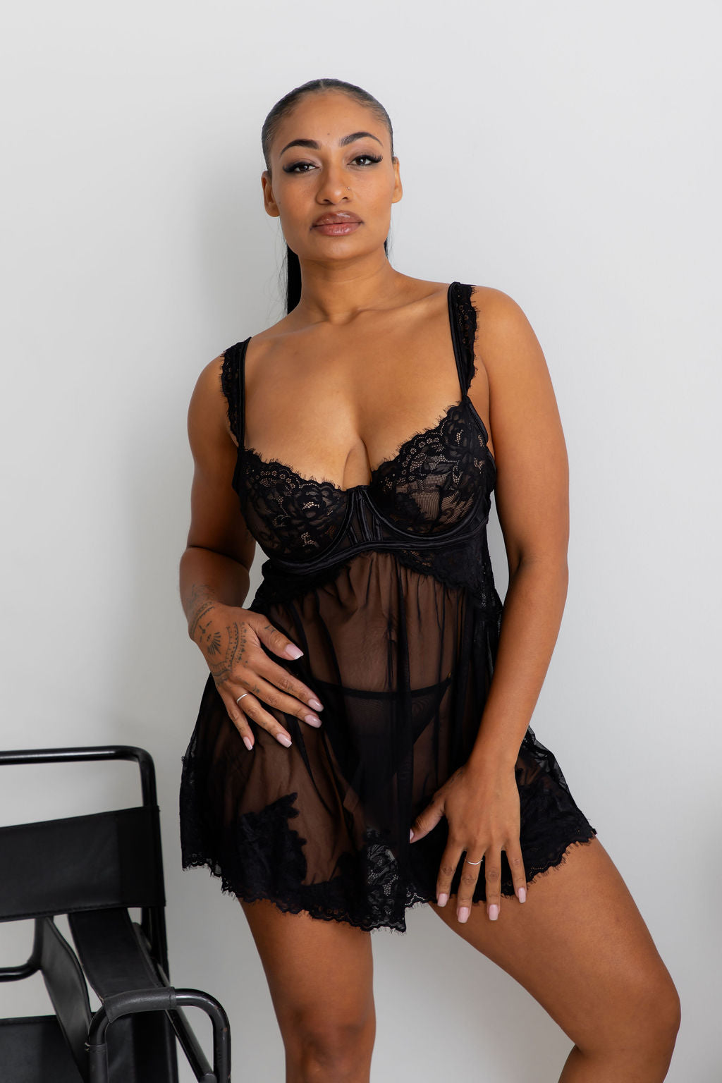 A woman modeling a black sheer lace babydoll lingerie chemise with a corset-style lace bodice, photographed against a plain light gray wall.