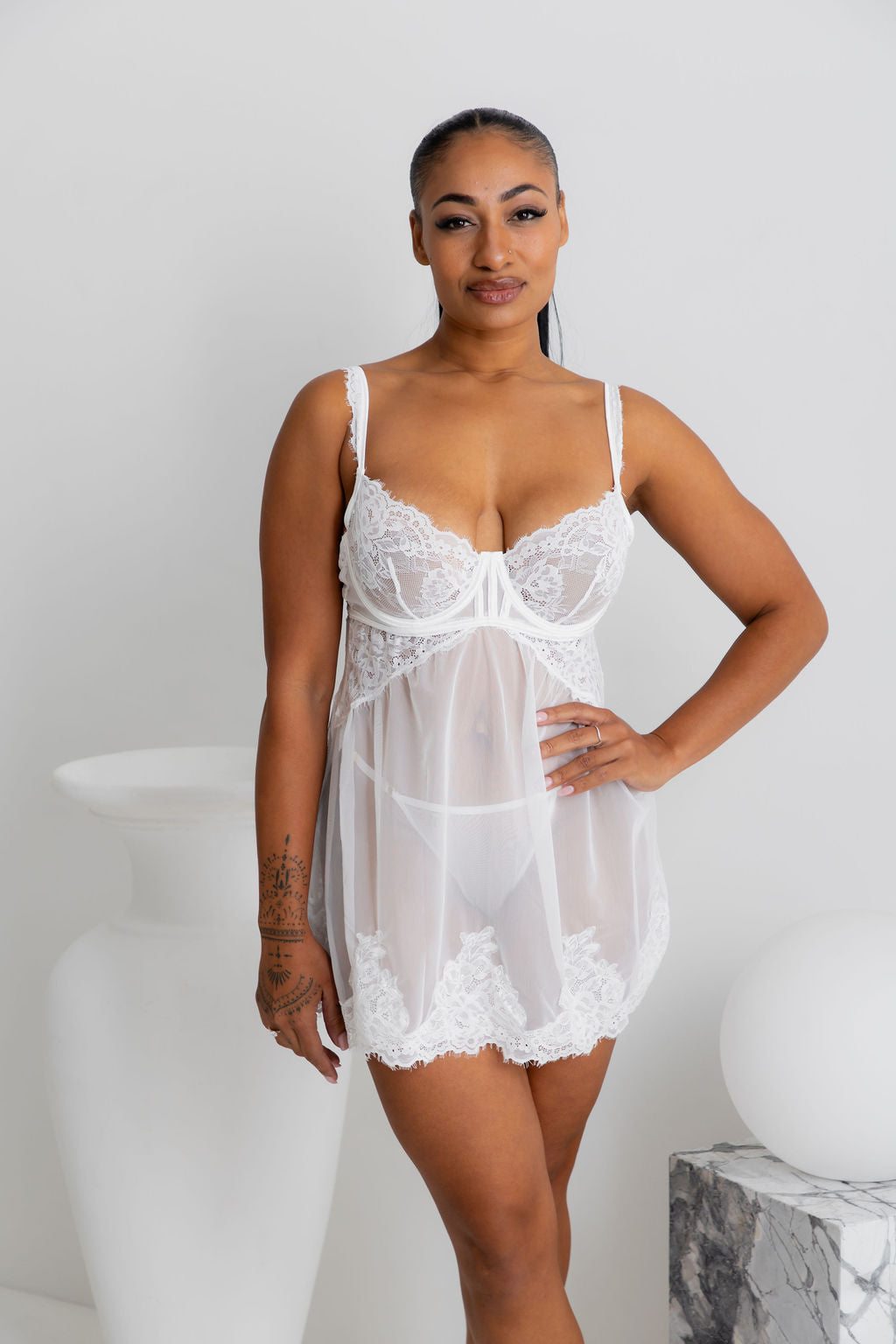 A woman modeling a white lace and sheer babydoll lingerie chemise in a minimalist studio setting with white decorative vases.