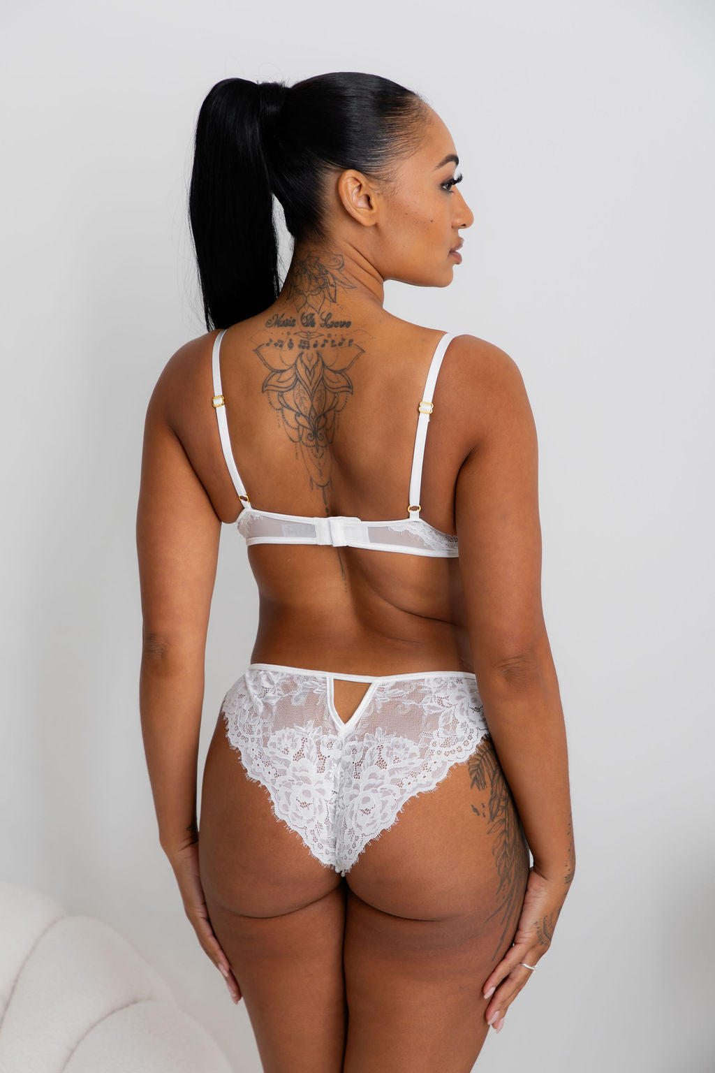 A woman shown from the back wearing a white lace lingerie set with a bra and matching panties, standing against a plain light gray wall.