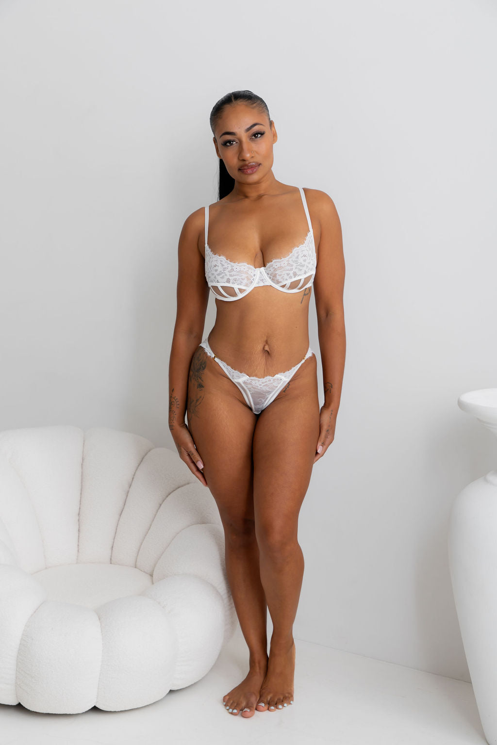 A woman modeling a white lace lingerie set consisting of a strappy underwire bra and matching high-cut briefs, standing in a bright white room next to a white shell-shaped chair.