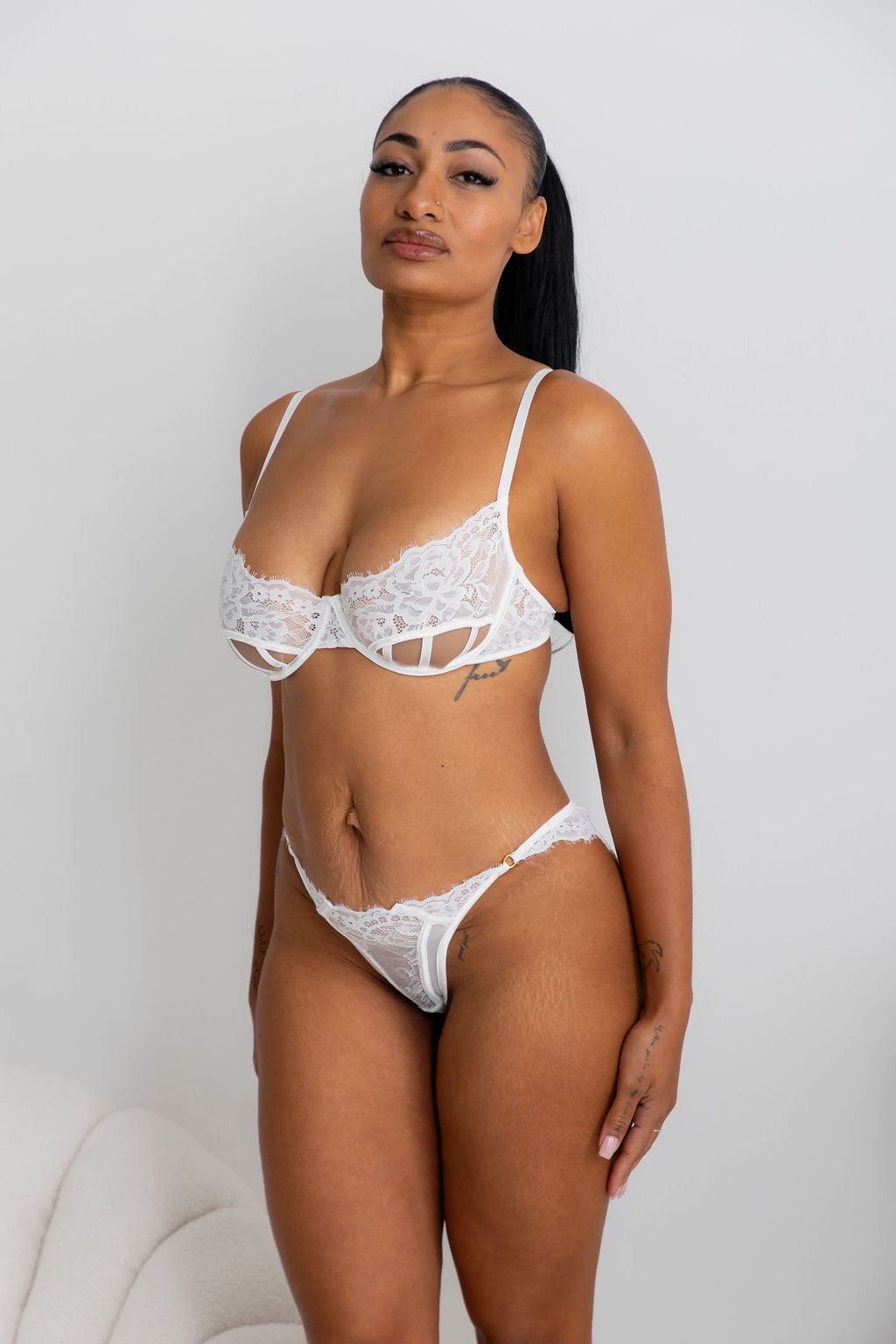A woman modeling a white lace lingerie set against a plain light gray wall. The set includes a strappy underwire bra and matching high-cut briefs with floral lace trim.