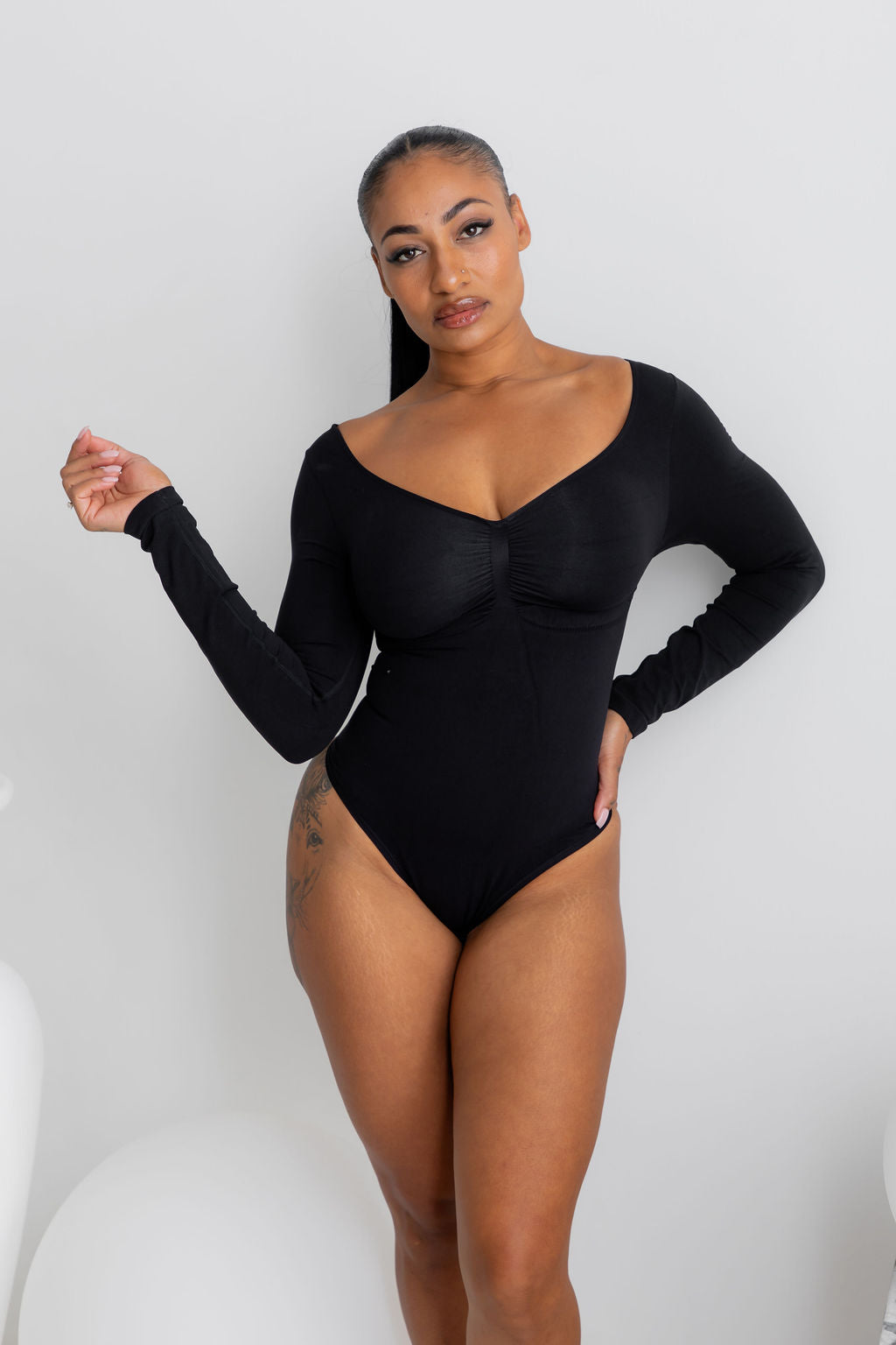 A woman modeling a black long-sleeve bodysuit with a deep scoop neckline and gathered bust detail, posed against a plain light gray studio background.