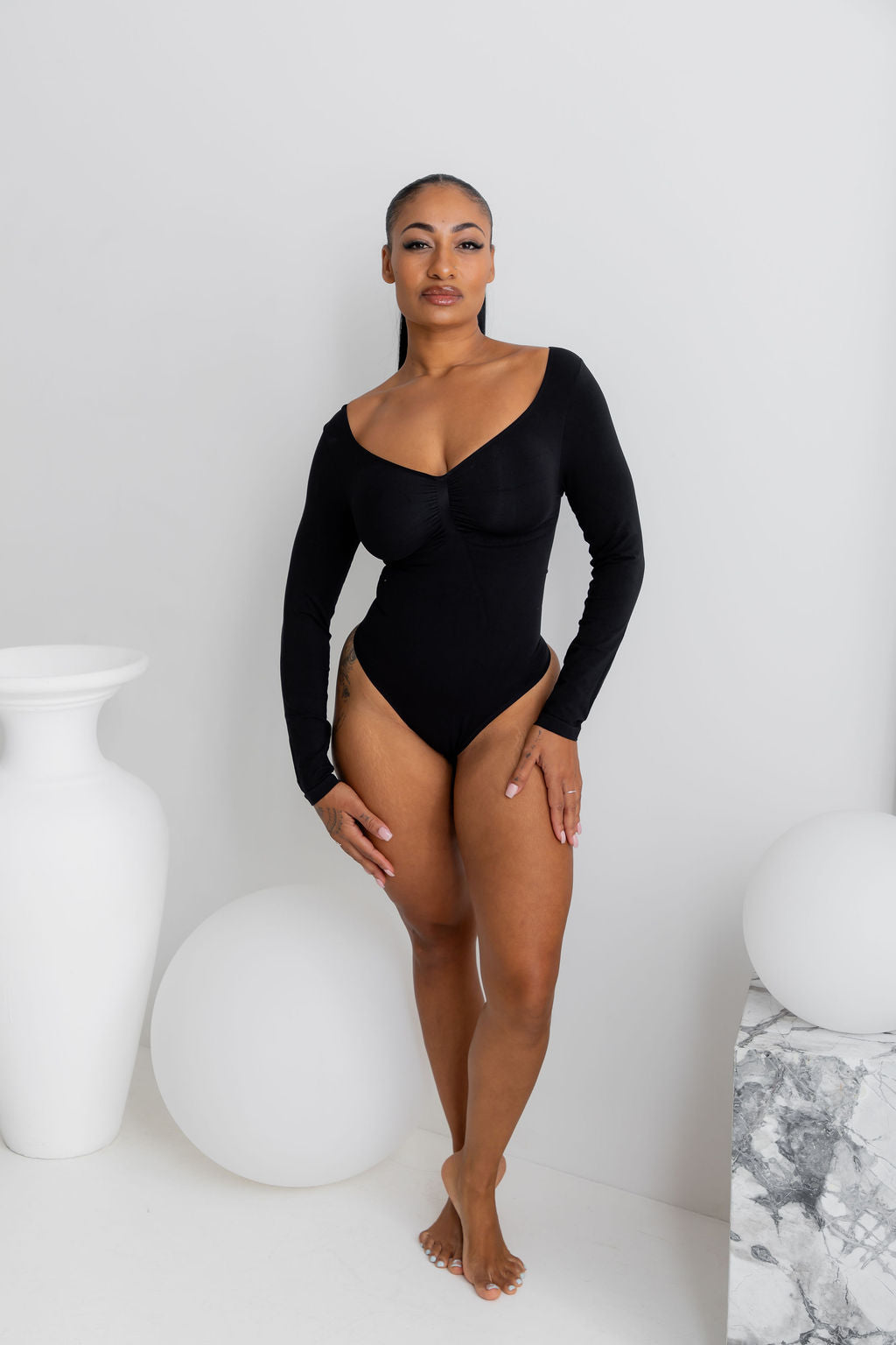 A woman modeling a black long-sleeve bodysuit with a deep scoop neckline in a minimalist studio setting with white decorative vases and spheres.