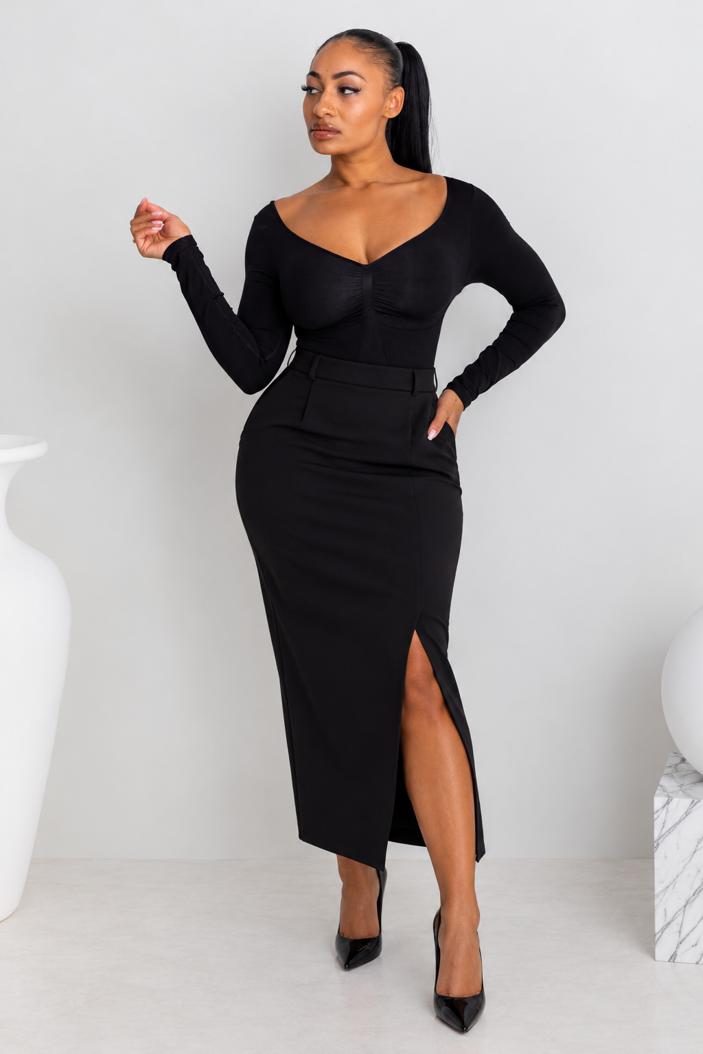 A woman modeling a black long-sleeve midi dress with a sweetheart neckline, defined waist, and a high front slit, standing in a minimalist studio setting.