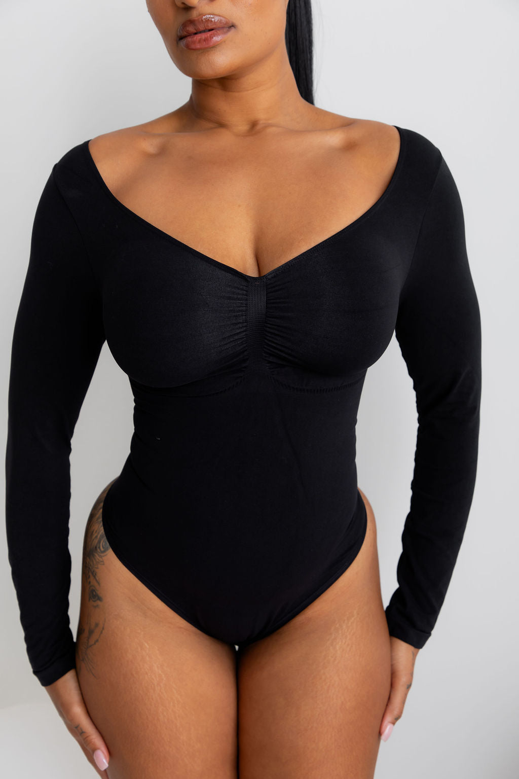 A woman wearing a black long-sleeve bodysuit with a deep scoop neckline and gathered bust detail, shown against a plain light gray studio background.