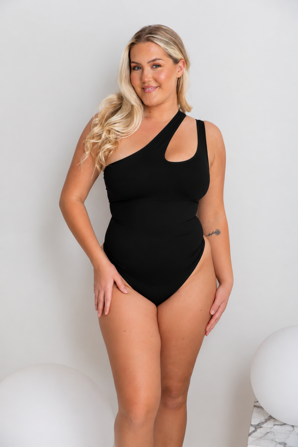 A blonde model wearing a black one-shoulder cutout swimsuit, standing against a light gray studio background with white decorative spheres.